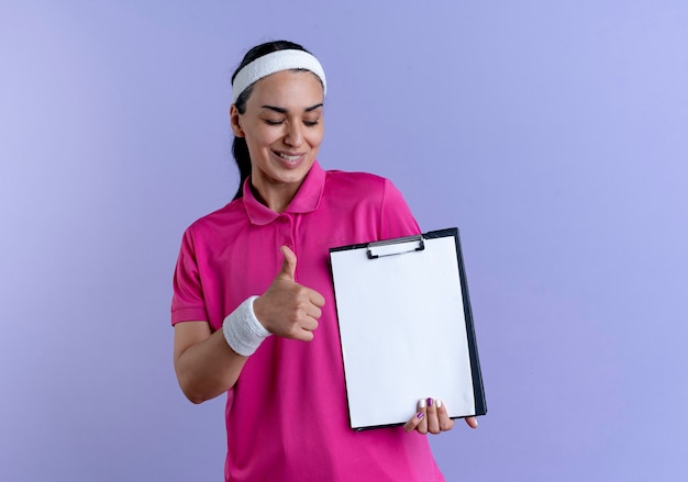 Sonriente joven mujer deportiva caucásica con diadema y muñequeras Thumbs up sosteniendo portapapeles aislado sobre fondo púrpura con espacio de copia