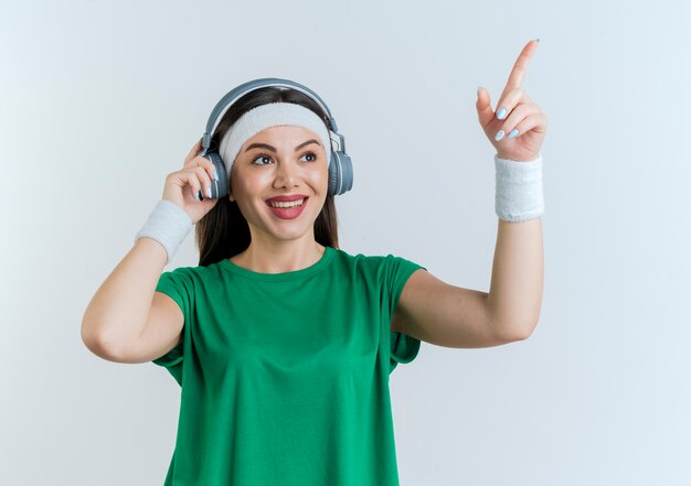 Sonriente joven mujer deportiva con diadema y muñequeras y auriculares mirando y apuntando al lado agarrando auriculares aislados en la pared blanca con espacio de copia