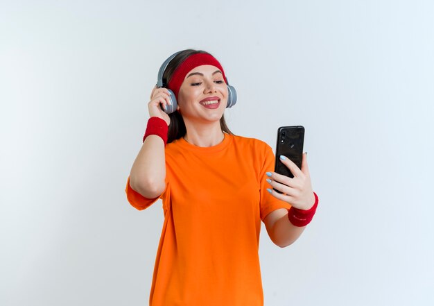 Sonriente joven mujer deportiva con diadema y muñequeras y auriculares sosteniendo y mirando el teléfono móvil agarrando auriculares aislados