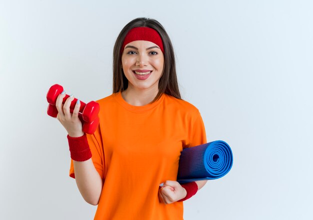 Sonriente joven mujer deportiva con diadema y muñequeras sosteniendo estera de yoga y pesas aisladas en la pared blanca con espacio de copia