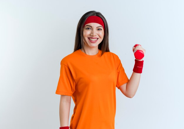 Sonriente joven mujer deportiva con diadema y muñequeras sosteniendo mancuernas aislado en la pared blanca con espacio de copia