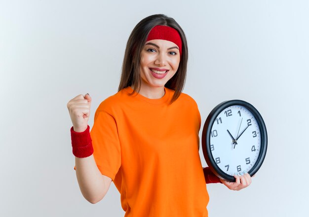 Sonriente joven mujer deportiva con diadema y muñequeras sosteniendo el reloj haciendo un gesto fuerte aislado en la pared blanca