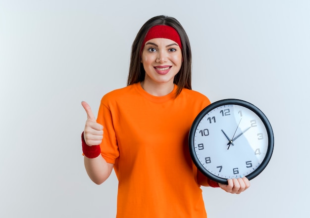 Sonriente joven mujer deportiva con diadema y muñequeras sosteniendo el reloj mostrando el pulgar hacia arriba aislado en la pared blanca con espacio de copia