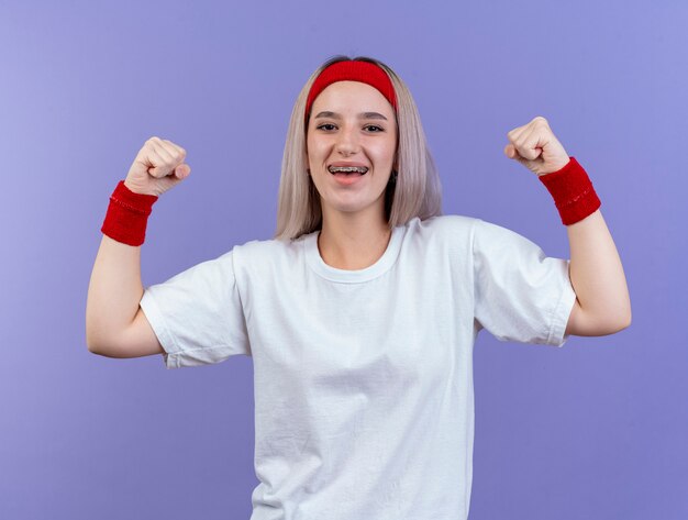 Sonriente joven mujer deportiva con tirantes con diadema y muñequeras tensa el bíceps aislado en la pared púrpura