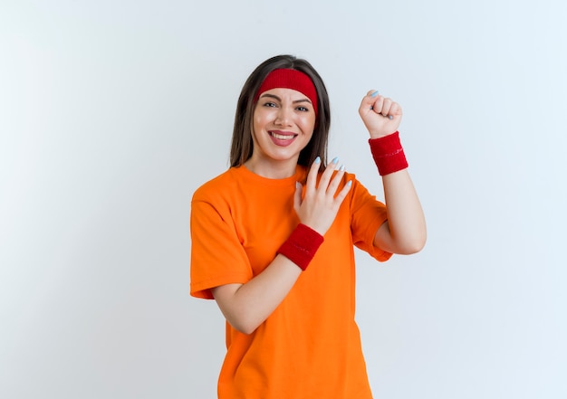 Sonriente joven mujer deportiva vistiendo diadema y muñequeras apretando el puño mirando manteniendo la mano en el aire aislado