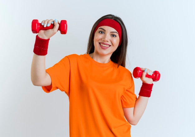 Sonriente joven mujer deportiva vistiendo diadema y muñequeras mirando sosteniendo pesas