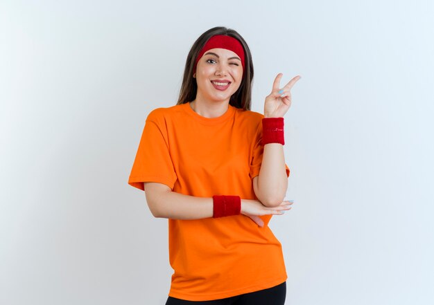 Sonriente joven mujer deportiva vistiendo diadema y muñequeras poniendo la mano debajo del codo guiñando un ojo haciendo el signo de la paz mirando aislado