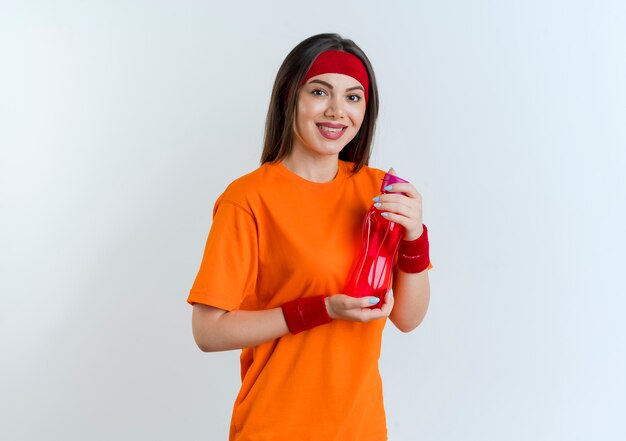 Sonriente joven mujer deportiva vistiendo diadema y muñequeras sosteniendo una botella de agua mirando aislado