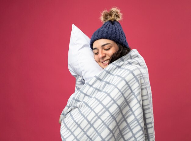 Sonriente joven mujer enferma con bata de invierno sombrero y bufanda envuelta en cuadros de pie en la vista de perfil abrazando la almohada con los ojos cerrados aislados en la pared rosa