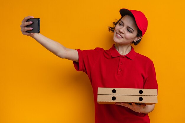 Sonriente joven mujer de entrega bonita sosteniendo cajas de pizza y tomando selfie en teléfono