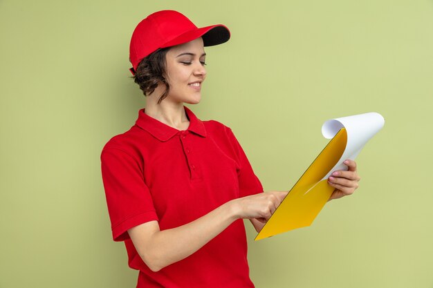 Sonriente joven mujer de entrega bonita sosteniendo y mirando el portapapeles