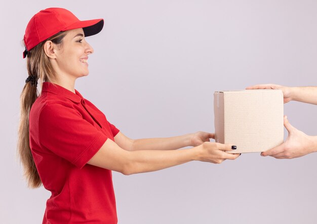 Sonriente joven mujer de entrega bonita en uniforme da caja de cartón a alguien aislado en la pared blanca