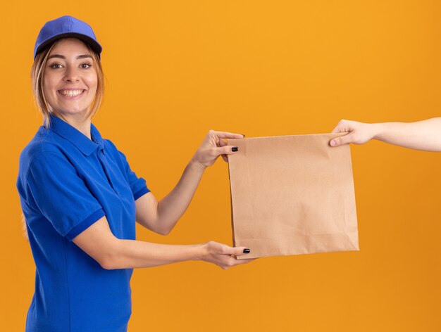 Sonriente joven mujer de entrega bonita en uniforme da paquete de papel a alguien que parece aislado