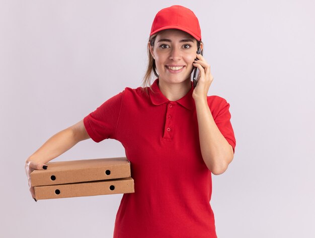 Sonriente joven mujer de entrega bonita en uniforme habla por teléfono y tiene cajas de pizza aisladas en la pared blanca