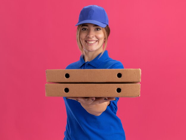 Sonriente joven mujer de entrega bonita en uniforme sostiene cajas de pizza y mira al frente aislado en la pared rosa