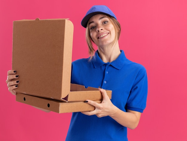Sonriente joven mujer de entrega bonita en uniforme tiene cajas de pizza aisladas en la pared rosa