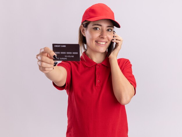 Sonriente joven mujer de entrega bonita en uniforme tiene tarjeta de crédito y habla por teléfono aislado en la pared blanca