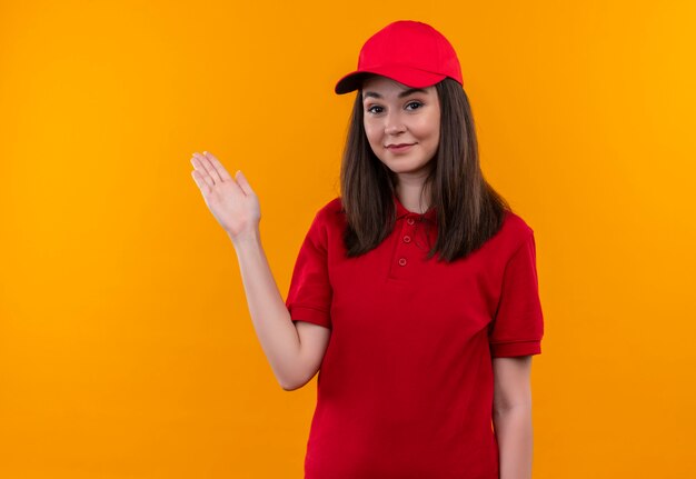 Sonriente joven mujer de entrega con camiseta roja con gorra roja apunta hacia el lado con la mano en la pared naranja aislada