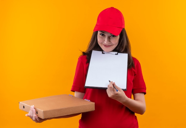 Sonriente joven mujer de entrega con camiseta roja en gorra roja con caja de pizza y portapapeles en pared naranja aislada