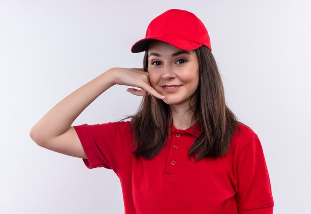 Sonriente joven mujer de entrega con camiseta roja con gorra roja hace una llamada con las manos en la pared blanca aislada