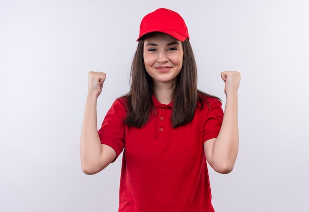 Sonriente joven mujer de entrega con camiseta roja en gorra roja levantó sus ajustes en la pared blanca aislada