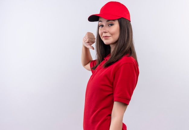 Sonriente joven mujer de entrega con camiseta roja en gorra roja mostrando los pulgares hacia abajo en la pared blanca aislada