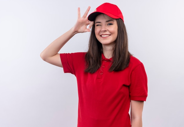 Sonriente joven mujer de entrega con camiseta roja en gorra roja parpadea en la pared blanca aislada