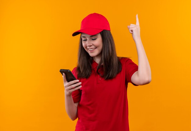 Sonriente joven mujer de entrega con camiseta roja en gorra roja y señala con el dedo hacia arriba en la pared amarilla aislada