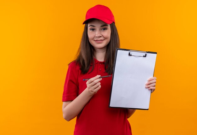 Sonriente joven mujer de entrega con camiseta roja en gorra roja sosteniendo una bandeja y portapapeles en la pared naranja aislada