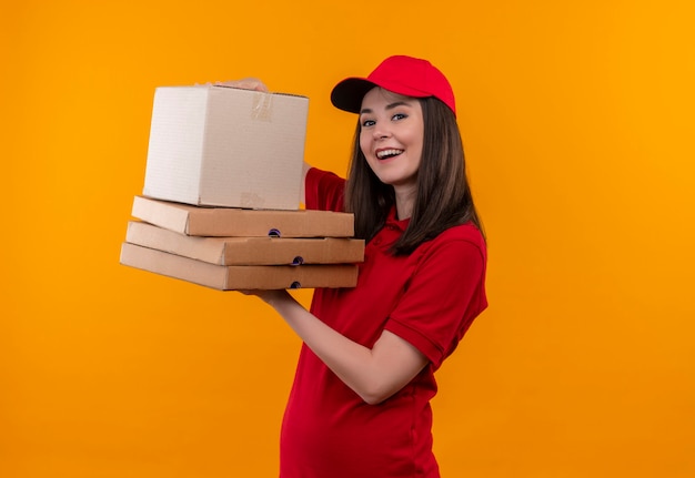 Sonriente joven mujer de entrega con camiseta roja en gorra roja sosteniendo una caja y caja de pizza en la pared amarilla aislada
