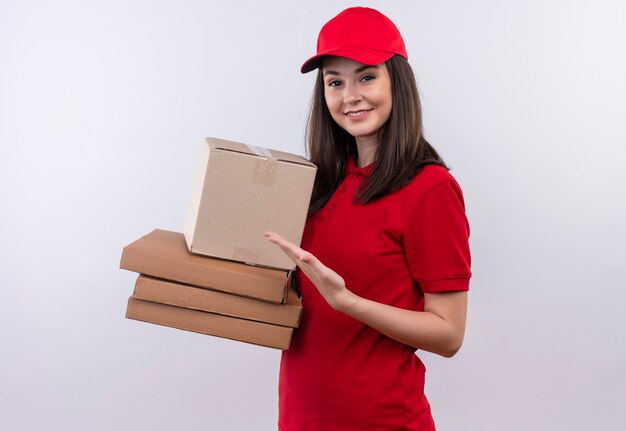 Sonriente joven mujer de entrega con camiseta roja en gorra roja sosteniendo una caja y caja de pizza en la pared blanca aislada