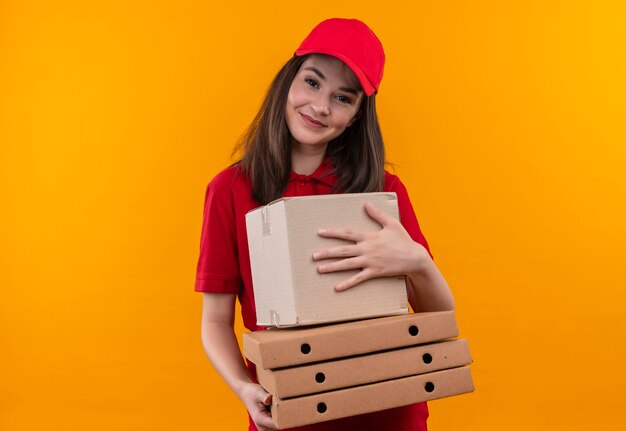 Foto gratuita sonriente joven mujer de entrega con camiseta roja en gorra roja sosteniendo una caja y caja de pizza en la pared naranja aislada