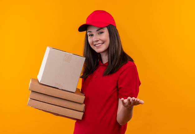 Sonriente joven mujer de entrega con camiseta roja en gorra roja sosteniendo una caja y caja de pizza en la pared naranja aislada