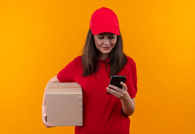 Sonriente joven mujer de entrega con camiseta roja en gorra roja sosteniendo una caja y hace una llamada en la pared naranja aislada