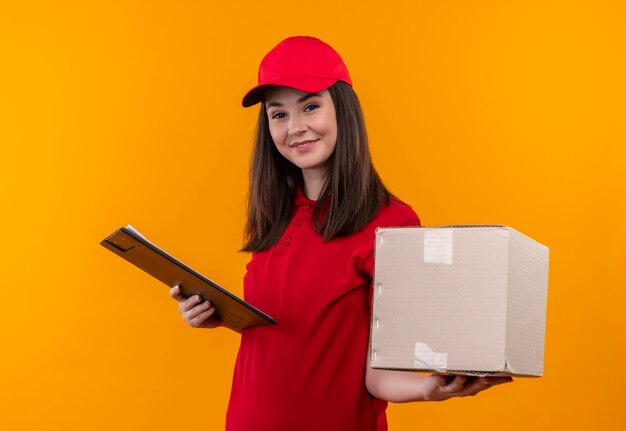 Sonriente joven mujer de entrega con camiseta roja en gorra roja sosteniendo una caja y portapapeles en la pared amarilla aislada