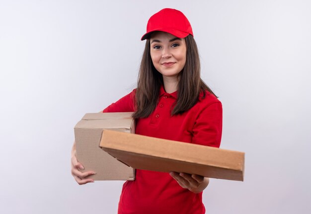 Sonriente joven mujer de entrega con camiseta roja en gorra roja sosteniendo una caja y sostiene la caja de pizza en la pared blanca aislada