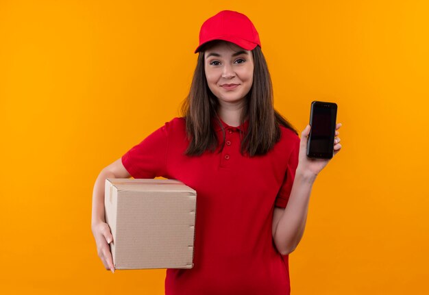Sonriente joven mujer de entrega con camiseta roja en gorra roja sosteniendo una caja y teléfono en la pared naranja aislada