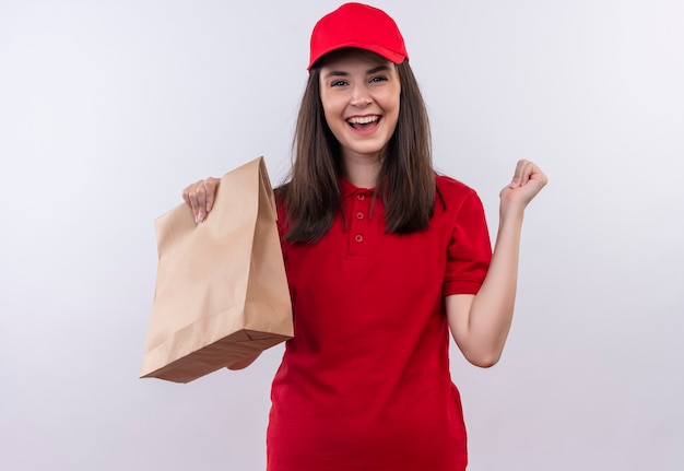 Sonriente joven mujer de entrega con camiseta roja en gorra roja sosteniendo el paquete en la pared blanca aislada