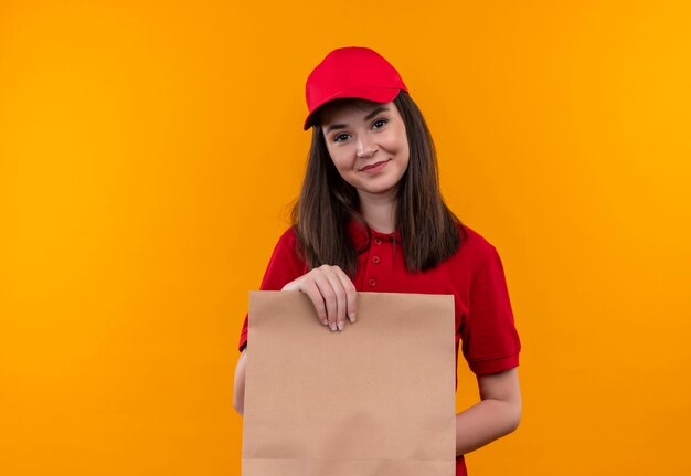 Sonriente joven mujer de entrega con camiseta roja en gorra roja sosteniendo el paquete en la pared naranja aislada