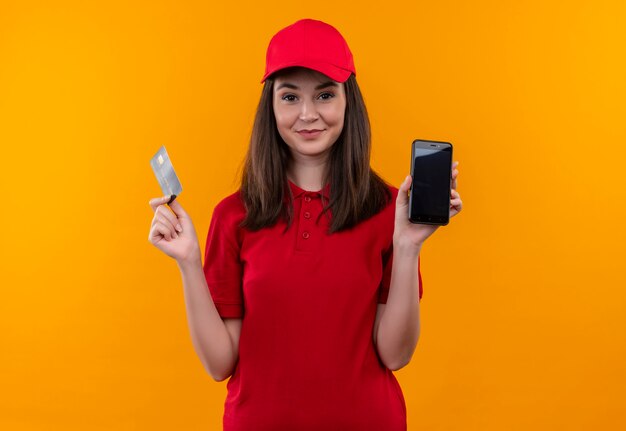 Sonriente joven mujer de entrega con camiseta roja en gorra roja sosteniendo la tarjeta y el teléfono en la pared naranja aislada