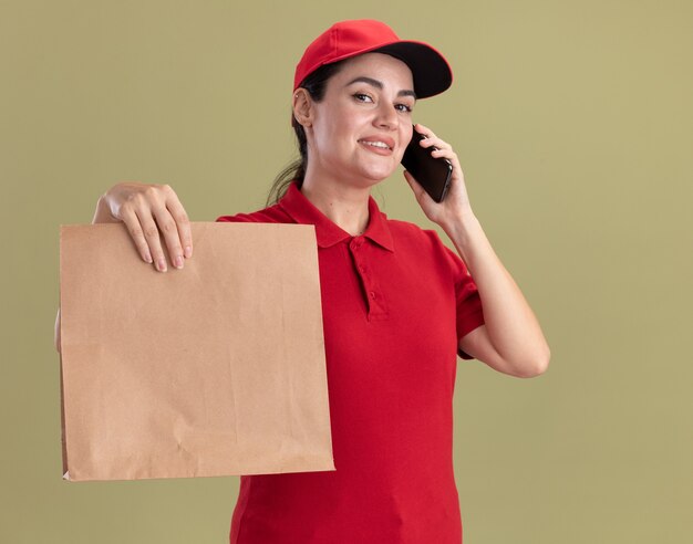 Sonriente joven mujer de entrega en uniforme y gorra hablando por teléfono estirando el paquete de papel hacia la cámara