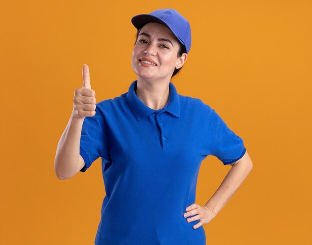 Sonriente joven mujer de entrega en uniforme y gorra manteniendo la mano en la cintura mostrando el pulgar hacia arriba