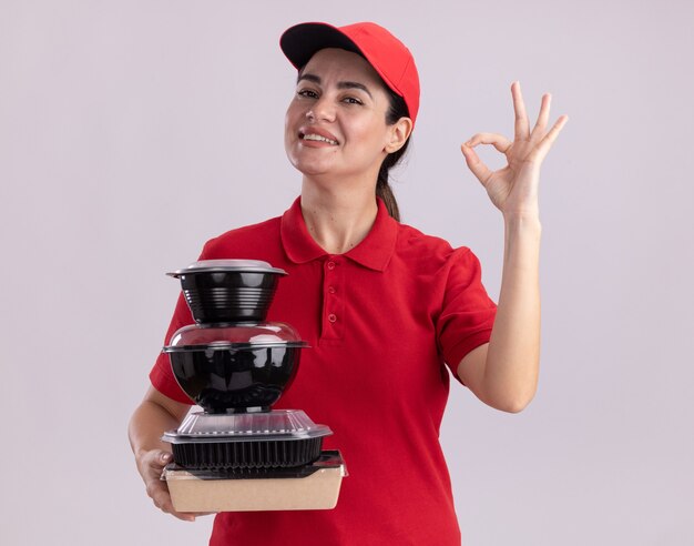 Sonriente joven mujer de entrega en uniforme y gorra con paquete de comida de papel y envases de comida haciendo el signo de ok