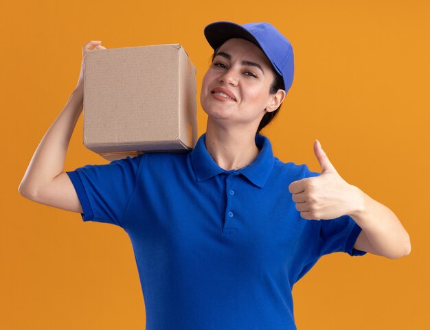 Sonriente joven mujer de entrega en uniforme y gorra sosteniendo cardbox en el hombro mostrando el pulgar hacia arriba aislado en la pared naranja