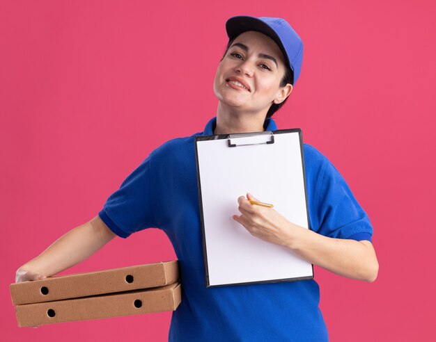 Sonriente joven mujer de entrega en uniforme y gorra sosteniendo paquetes de pizza apuntando con lápiz en el portapapeles