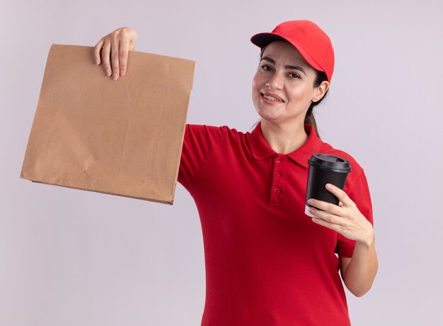 Sonriente joven mujer de entrega en uniforme y gorra sosteniendo una taza de café de plástico y un paquete de papel