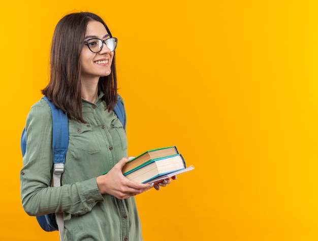 Sonriente joven mujer de la escuela con mochila con gafas sosteniendo libros