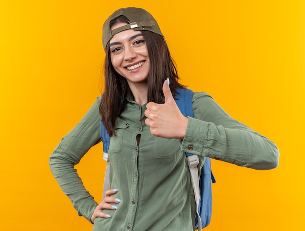 Sonriente joven mujer de la escuela con mochila con gorra mostrando el pulgar hacia arriba poniendo la mano en la cadera