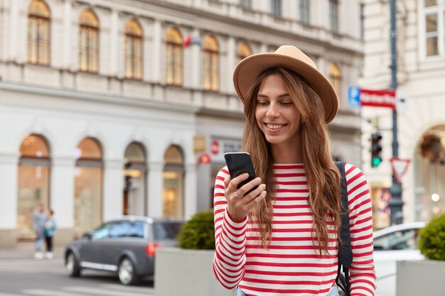 Sonriente joven mujer europea disfruta del tiempo de recreación, mira videos interesantes en el sitio web, usa un elegante sombrero y un suéter a rayas