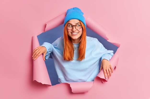 Sonriente joven mujer europea jengibre con expresión alegre mira hacia otro lado felizmente expresa gran interés viste sombrero azul y jersey de manga larga.